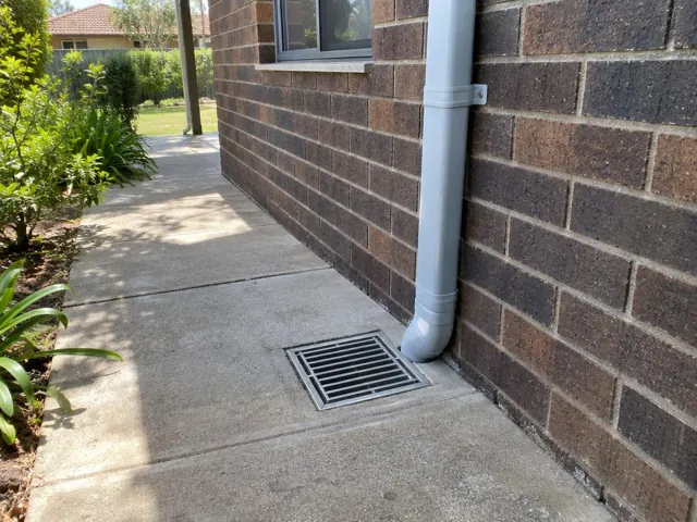 Outdoor gully grate beside a brick house wall with dry concrete paving in Carlingford