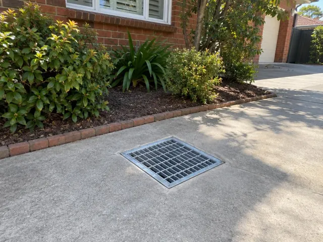 Metal stormwater grate set flush in a concrete driveway beside garden beds in Epping